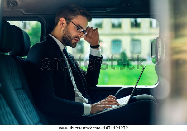 Side\
view of confident young man in full suit working using laptop while\
sitting in the car being fully concentrate on\
work