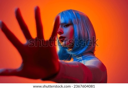 Side view of confident young woman in blue wig with red lips covering camera with hand against orange background