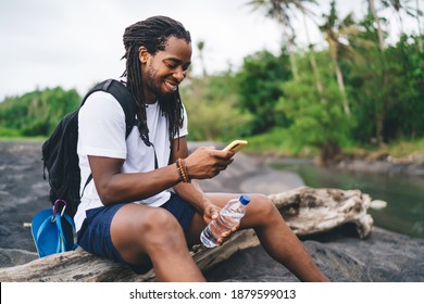 Side View Content African American Male Hiker Sitting On Dry Tree Trunk And Browsing Mobile Phone While Resting In Tropical Forest