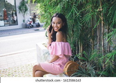 Side view of content charming young Asian woman in pink off-shoulder dress speaking on smartphone while sitting on parapet against wall with green tropical foliage