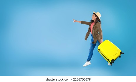 Side view of cool young black lady running and jumping with bright suitcase, pointing finger at empty space over blue studio background, banner design. Summertime vacation, travelling concept