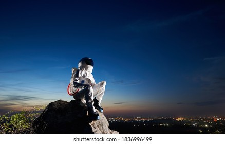 Side View Of Cosmonaut Sitting On Top Of Rocky Hill And Looking At Beautiful Night Sky. Astronaut Wearing Space Suit And Helmet. Concept Of Astronautics And Human Space Exploration.