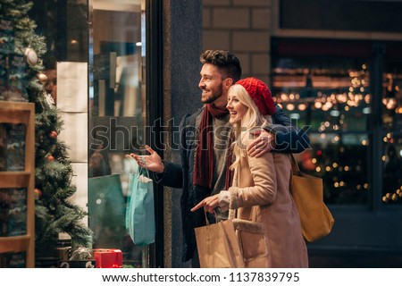 Side view of a couple doing some window shopping at christmas. The mid adult male has his arm around the mid adult female and they are talking about wats in the window.