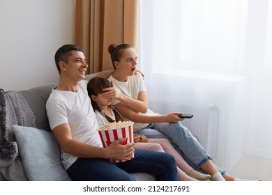 Side View Of Couple Sitting On Sofa And Watching Movie Together, Mother Closed Daughter's Eyes And Looking With Open Mouth, Father Smiling, Family Spending Time Together.