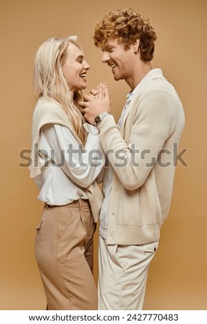 side view of couple in trendy light-colored clothes holding hands and laughing on beige backdrop