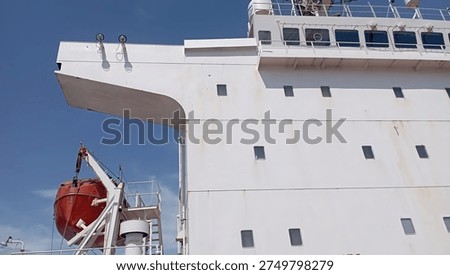 Side view of Crew accommodation on board a ship's white superstructure featuring an orange emergency lifeboat and navigation bridge against a blue sky.