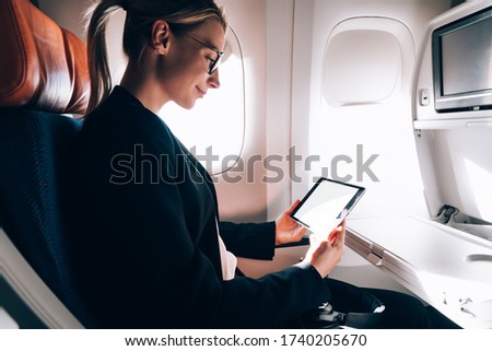 Side view of crop delighted female in elegant outfit and eyeglasses reading e book on tablet and chilling during flight while sitting near window in aircraft