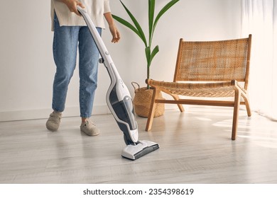Side view of crop faceless housewife in casual outfit and slippers cleaning laminate floor with modern upright vacuum cleaner in minimalist light apartment