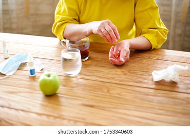 Side View Of Crop Middle Aged Female Taking Pills While Sitting At Table With Cup Of Tea And Protective Mask At Home