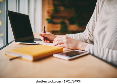 Side View Of Crop Person Sitting At Table With Laptop And Smartphone While Writing Notes In Notepad During Workday In Modern Office