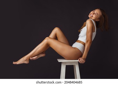 Side view cropped portrait of young beautiful woman sitting on white chair and laughing on dark brown background. Perfect body shape. Natural beauty. Concept of health, beauty, youth, spa, fitness, ad