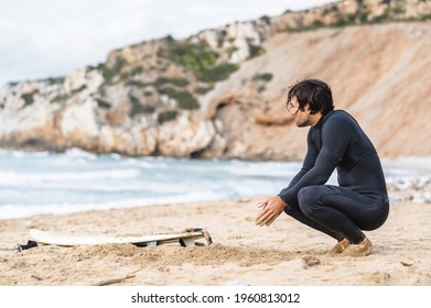 Side view of crouched wet man wearing diving suit. Male on the beach with his surfboard resting and looking at the sea after surfing. - Powered by Shutterstock