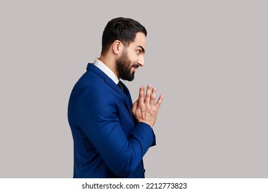Side view of cunning bearded man clasping hands and planning evil tricky prank or scheming, cheating with sly smile, wearing official style suit. Indoor studio shot isolated on gray background.