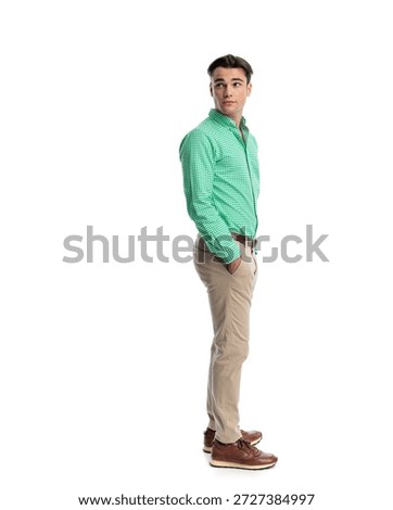 Side view of curious man standing with hands in pockets looking back over shoulder while wearing green checkered shirt and beige pants isolated on white background
