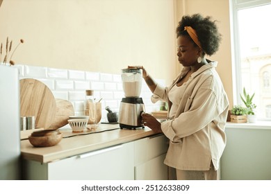 Side view of curly Black woman using food blender mixing ingredients for milk smoothie while cooking healthy breakfast at beige sunlit kitchen, copy space - Powered by Shutterstock