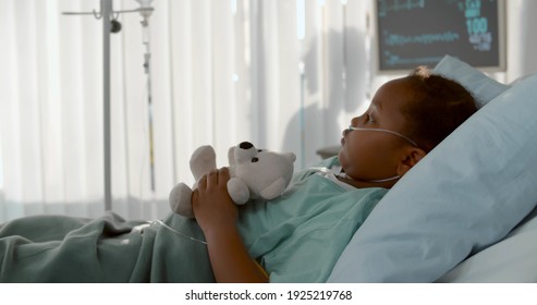 Side view of cute african girl lying in hospital bed with white teddy bear while doctors working behind curtain. Kid patient resting in pediatrics department - Powered by Shutterstock