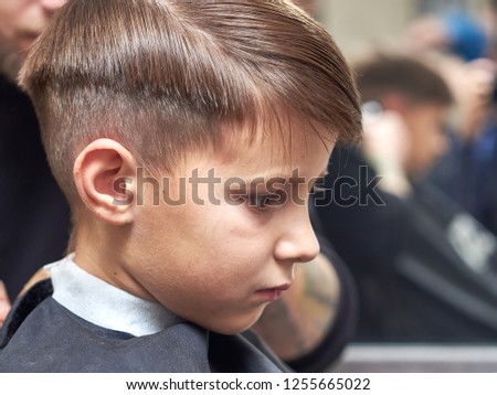 Side view of cute boy getting hairstyle by hairdresser in barbershop.