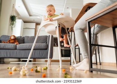 Side view of cute caucasian baby sitting in high chair with piece of vegetable looking with interest on messy and dirty floor covered with broccoli, carrot and cauliflower. Baby feeding concept