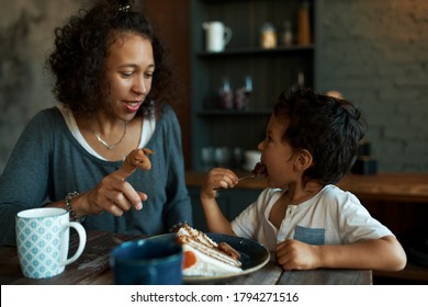 Side view of cute little boy eating cake at kitchen table with his young mother sitting next to him playing with finger puppet. Attractive Latin woman entertaining her baby son during lunch
