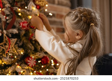 Side view cute little child girl in white warm sweater is decorating Christmas tree in living room at home. Merry Christmas and Happy New year winter holidays. High quality photo - Powered by Shutterstock