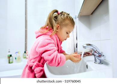 Side view of cute little girl with ponytail in pink bathrobe washing her hands. Copyspace.