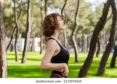 Side View Of Cute Sportive Woman Wearing Black Sports Bra Standing On City Park, Outdoors Enjoying Stress Free Mindful Moment, Doing Yoga Relaxation Exercises, Closed Eyes, Meditating Nature.