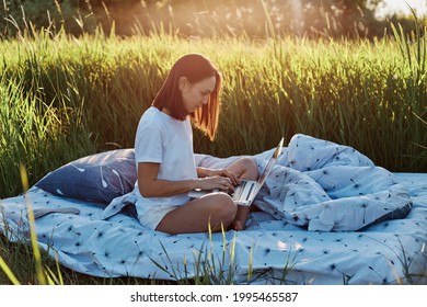 Side View Of Dark Haired Woman Working Outdoor, Female In Casual Attire Holding Lap Top On Knees, Freelancer Doing Her Job In Green Meadow On Sunset, Enjoying Nature.