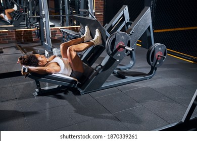 Side view of a dark-haired strong well-built lady athlete using the incline leg press machine
