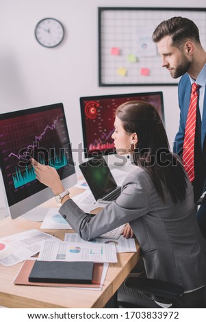 Side view of data analyst pointing with hand at graphs on computer monitor to colleague in office