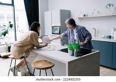 Side view of daughter sitting on bar stool showing planner to senior mother pointing at note discussing plans while spending time in contemporary kitchen - Powered by Shutterstock