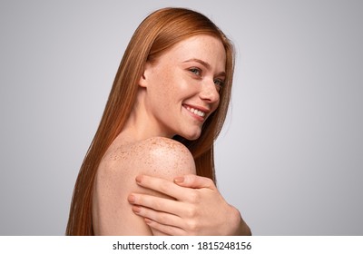 Side View Of Delighted Ginger Lay Smiling And Looking Away While Embracing Freckled Shoulder During Skin Care Routine Against Gray Background