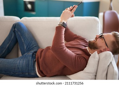Side view of delighted male in glasses lying on couch and messaging on social media via mobile phone at home