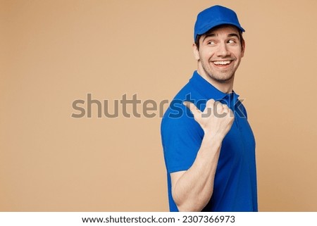 Side view delivery guy employee man wearing blue cap t-shirt uniform workwear work as dealer courier point thumb finger aside indicate on area isolated on plain light beige background. Service concept