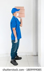 Side view of delivery man wearing face mask and gloves while carrying a stack of packages and standing near an opened door of customer house