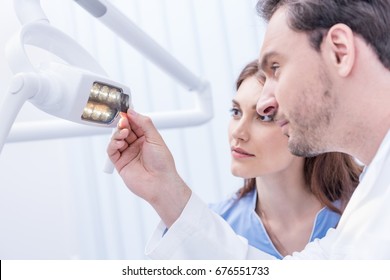 Side View Of Dentists Looking At Xray Picture Of Teeth In Hand Against Dental Lamp 