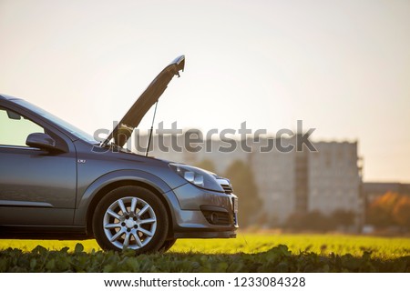 Side view detail of car with open hood on empty gravel field road on blurred apartment building and clear bright sky copy space background. Transportation, vehicles problems and breakdowns concept.