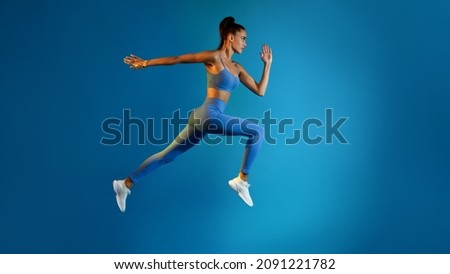 Side View Of Determined Young Sportswoman Jumping Posing In Mid-Air Looking Aside On Blue Studio Background. Serious Fitness Female Running In Air. Panorama, Full Length