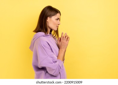 Side view of devious cunning young woman clasping hands and smirking mysteriously, scheming cheats, evil prank, wearing purple hoodie. Indoor studio shot isolated on yellow background.