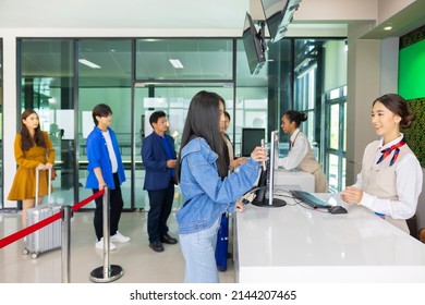 Side View Of Different Ages And Nationalities Passengers Standing In Queue To Check In At Airport Terminal. Young Asian Woman Showing Flight Ticket To Staff On Phone And Waiting For Her Boarding Pass.