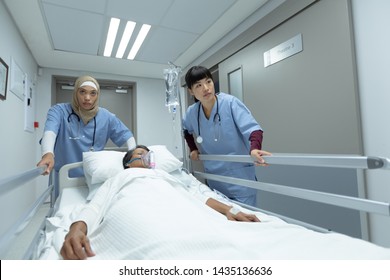 Side View Of Diverse Female Doctors Pushing Emergency Stretcher Bed In Corridor At Hospital. Mixed-race Female Patient With Oxygen Mask Lying In Stretcher Bed.