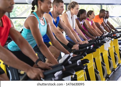 Side View Of Diverse Fit People Exercising On Exercise Bike In Fitness Center. Bright Modern Gym With Fit Healthy People Working Out And Training At Spin Class