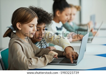 Side view at diverse group of children sitting in row at school classroom and using laptops