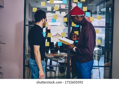 Side View Of Diverse Male Coworkers Standing Near Glass Wall With Sticky Notes And Discussing Diagrams While Working On Marketing Project In Modern Office