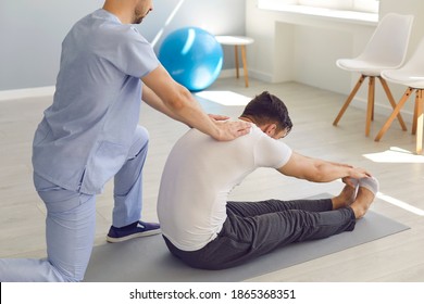 Side View Doctor Helps The Patient To Do Stretching Exercises And Yoga After The Injury. Man Sitting On A Sports Mat Stretches His Arms To His Toes. Concept Of Professional Medical Care.