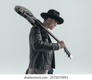 side view of dramatic young singer leaving with the guitar on his shoulder in front of grey background