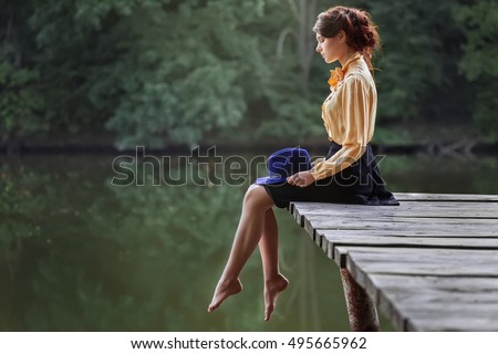 Side view of dreaming girl sitting on jetty forest and river on background. Sad girl depressed on bridge by the lake
