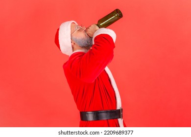Side view of drunk elderly man with gray beard wearing santa claus costume celebrating winter holidays with alcohol, drinking beer with closed eyes. Indoor studio shot isolated on red background.
