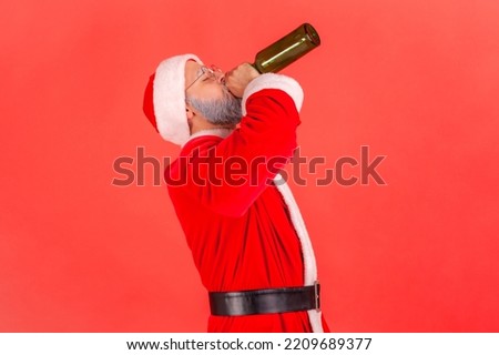 Side view of drunk elderly man with gray beard wearing santa claus costume celebrating winter holidays with alcohol, drinking beer with closed eyes. Indoor studio shot isolated on red background.