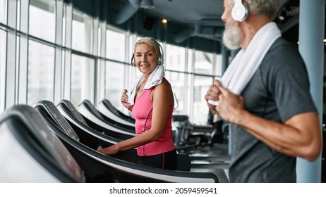 Side View Elder Smiling Woman Working Out On Treadmill, Looking At Camera, Selective Focus. Senior Man In Gray T-shirt, Blured. Elder Couple Enjoying The Moment. Modern Gym On Backgrund.