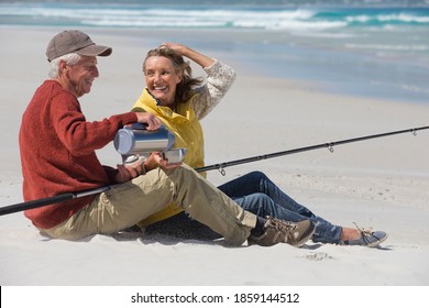 Side view of an elderly couple with a fishing rod sitting on a sunny beach and enjoying coffee. - Powered by Shutterstock